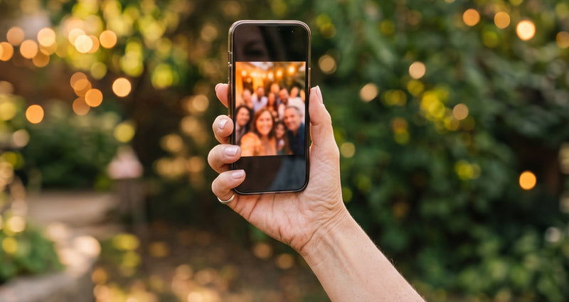 A hand holding up a phone showing a warm group photo, string lights and greenery behind it.